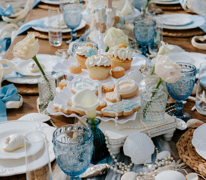 Coastal-chic beachside dessert table with pastel cupcakes and iced cookies on a tiered stand, blue goblets, pale roses, seashells and pearl garlands on woven chargers by the sand