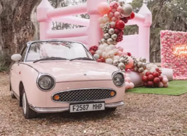 Pink vintage-style convertible parked by a pastel pink inflatable bounce house with a cascading pink, red and metallic balloon garland at an outdoor woodland party
