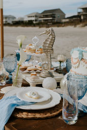 Seaside dessert table on a sandy beach with a metallic seahorse centerpiece, pastel shell‑decor cake and cupcakes, blue glass goblets, calla lilies and scattered seashells, beachfront houses blurred in the background.