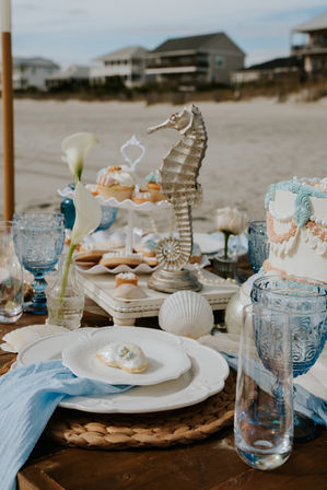 Seaside dessert table on a sandy beach with a metallic seahorse centerpiece, pastel shell‑decor cake and cupcakes, blue glass goblets, calla lilies and scattered seashells, beachfront houses blurred in the background.