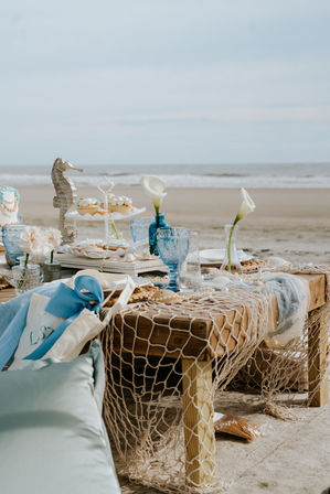 Coastal picnic setup on a sandy beach with ocean in the background — nautical decor, blue glassware, calla lilies, a seahorse ornament, cupcakes and scattered seashells.