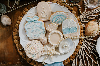 Coastal-inspired iced sugar cookies in seafoam blues and creams on a white plate surrounded by seashells, a starfish, pearl beads and braided placemat on a wooden table