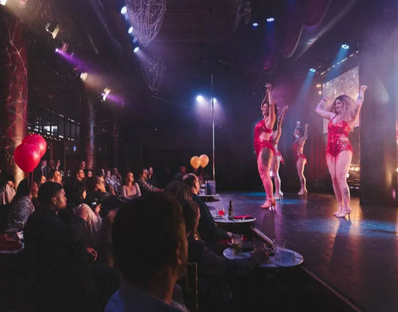 Cabaret nightclub scene with burlesque dancers in red sequined costumes performing under purple-pink stage lights as a seated audience at small tables watches, balloons and drinks visible.