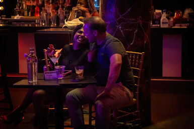 Couple laughing and leaning in over cocktails at a dimly lit bar table with a 'Reserved' sign, purple neon mood lighting and bottles on the back bar.