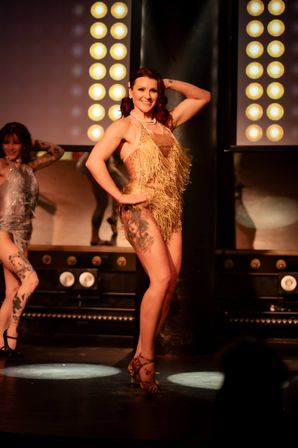 Burlesque performer in a gold fringe costume and pearl necklace posing on a cabaret stage under vintage round stage lights, showing a leg tattoo with a backup dancer in silver visible
