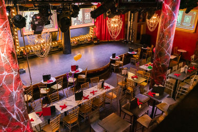 Empty cabaret-style dinner theater set for a burlesque show with a curved stage, red curtains, sparkling chandeliers, gold chairs and tables arranged with menus and red napkins.