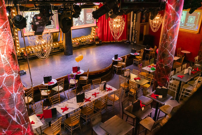 Empty cabaret-style dinner theater set for a burlesque show with a curved stage, red curtains, sparkling chandeliers, gold chairs and tables arranged with menus and red napkins.