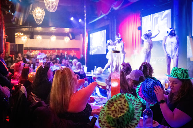 Packed nightclub cabaret with a lively seated crowd in neon animal-print hats and tiaras watching sparkly-costumed dancers perform on a colorful, chandelier-lit stage.