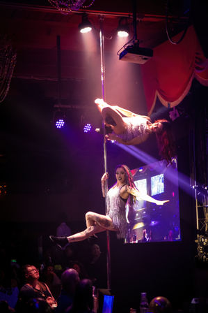 Two pole dancers in silver fringe costumes performing aerial tricks on a nightclub cabaret stage under purple spotlights, watched by a seated audience.