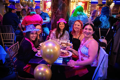 Group of five friends smiling at a decorated table in a colorful indoor nightlife venue, wearing party hats, tiaras and feather boas with gold balloons and plates of food — lively birthday or bachelorette celebration.