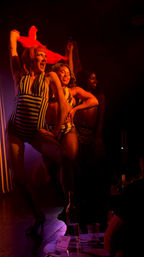 Three sassy drag performers in striped costumes and fishnet stockings strike a choreographed pose on a dim nightclub/cabaret stage bathed in red light, with audience tables and drinks visible in the foreground.