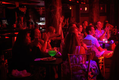 Audience at an indoor nightlife venue bathed in red stage lighting, seated at small tables and clapping along to a live show, with drinks and a glowing green cocktail on a foreground table.