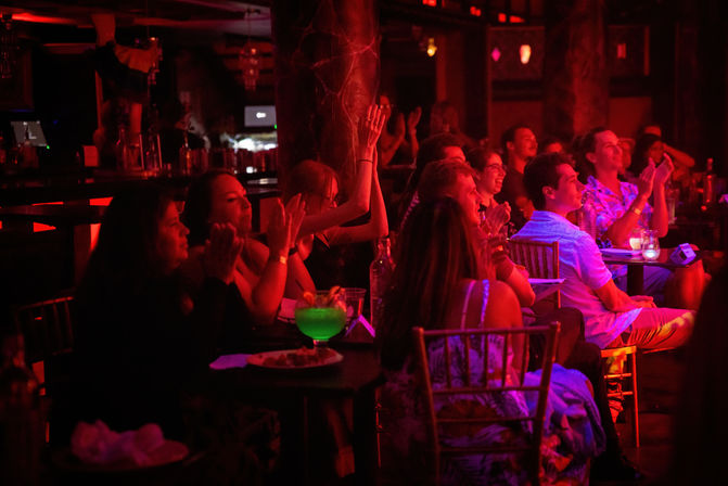 Audience at an indoor nightlife venue bathed in red stage lighting, seated at small tables and clapping along to a live show, with drinks and a glowing green cocktail on a foreground table.
