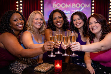 Five women smiling and toasting with champagne flutes in a lively lounge or nightclub, marquee lights and purple stage backdrop, red votive candle and sparkly clutch on the table — girls' night out celebration.