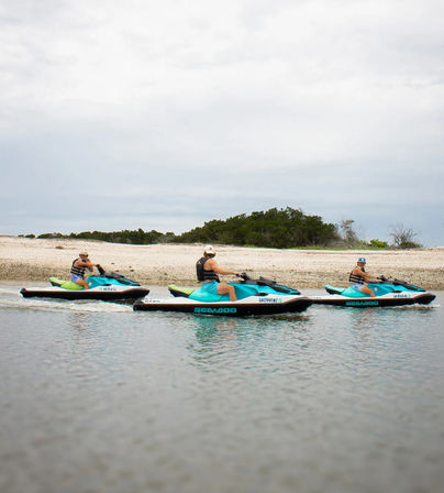 Three people on teal jet skis riding in calm coastal waters past a sandy beach and low green shrubs under an overcast sky.