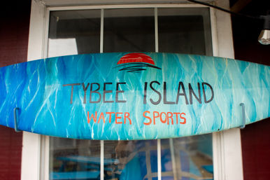 Blue-green painted surfboard sign with a red sun graphic and the words "Water Sports," mounted above a window at a beachside shop.