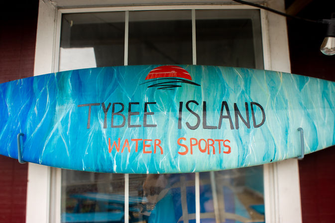 Blue-green painted surfboard sign with a red sun graphic and the words "Water Sports," mounted above a window at a beachside shop.