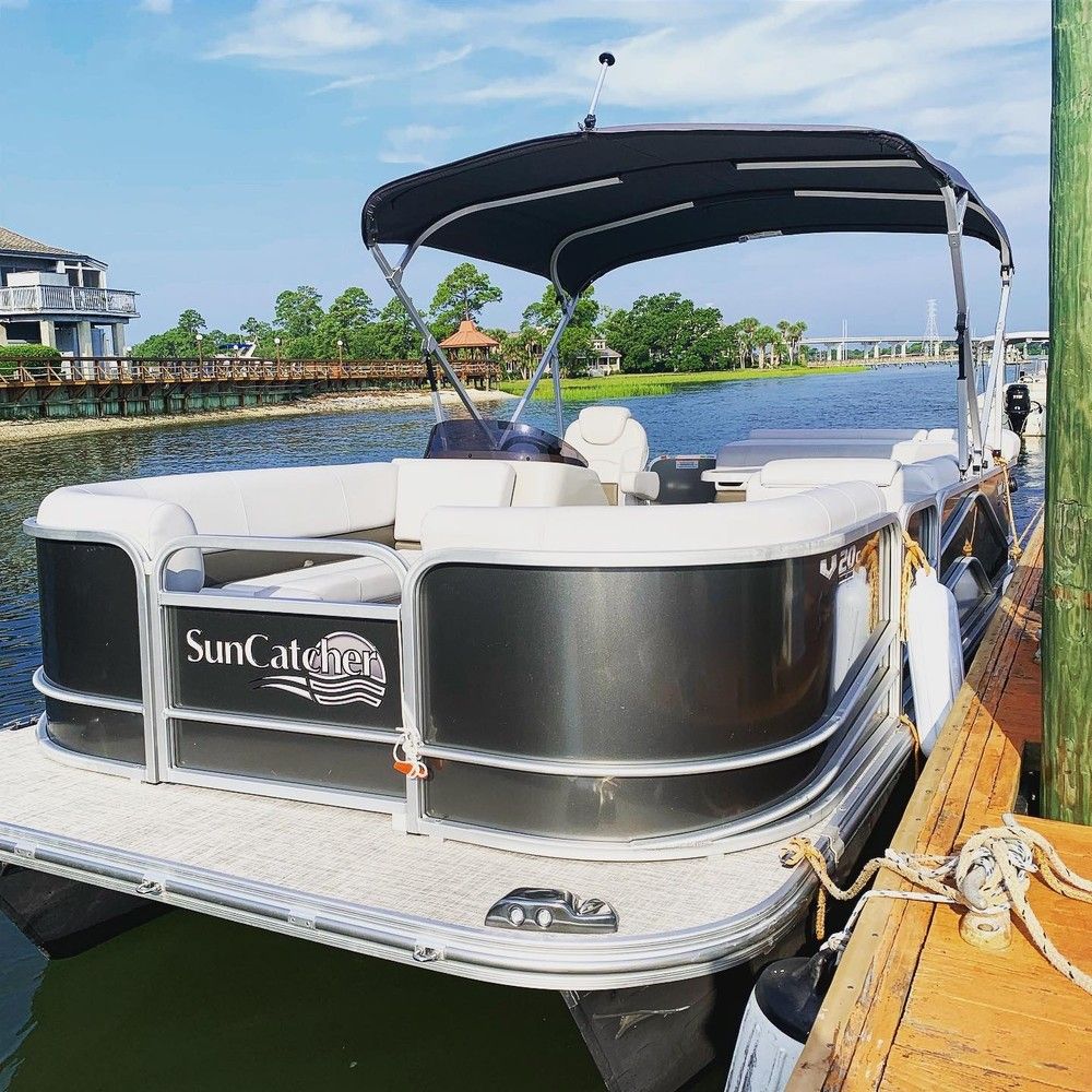 Sleek gray-and-white pontoon boat with black bimini top moored at a wooden dock on a calm waterfront river, with homes, a boardwalk and green trees under a blue summer sky.