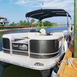Sleek gray-and-white pontoon boat with black bimini top moored at a wooden dock on a calm waterfront river, with homes, a boardwalk and green trees under a blue summer sky.
