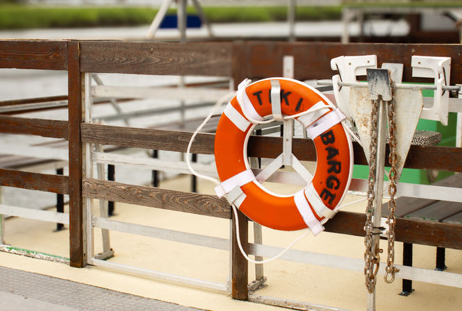 Bright orange lifebuoy with white straps hung on a weathered wooden dock railing, rusty anchor chain and metal fittings nearby, blurred marina water in background