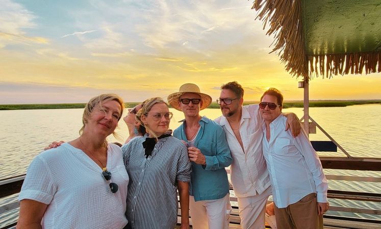Group of adults posing and smiling on a wooden waterfront dock under a thatched roof, golden sunset reflecting across a calm marsh inlet