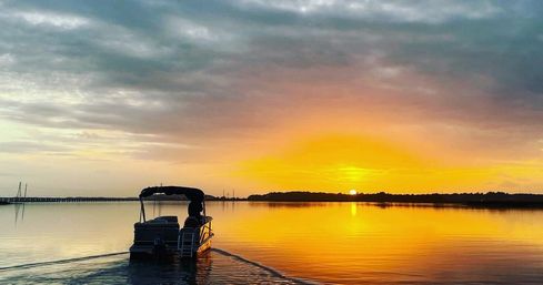 Pontoon boat cruising across a mirror-calm bay toward a golden sunset, orange sky reflected on glassy water with a distant pier