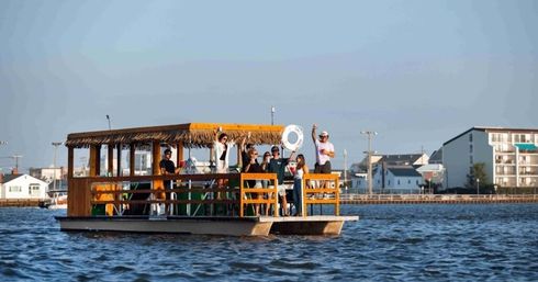 Group of people enjoying a sunny summer cruise on a wooden tiki-style pontoon boat near a coastal waterfront with low-rise buildings and calm blue water