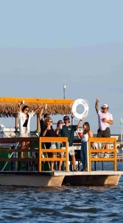 Group of people enjoying a sunny summer cruise on a wooden tiki-style pontoon boat near a coastal waterfront with low-rise buildings and calm blue water