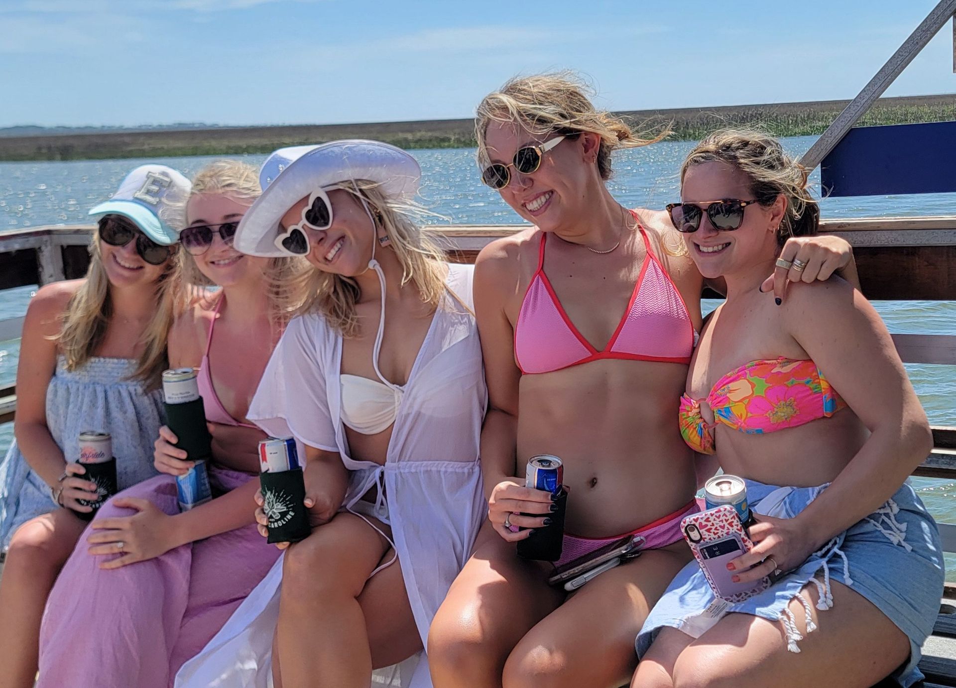 Five friends in bikinis and sunglasses smiling on a wooden boat across a sunny marshy lake, holding canned drinks and wearing summer hats and cover-ups