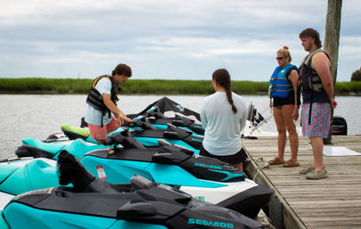 Four adults in life jackets at a wooden dock checking turquoise jet skis (personal watercraft), ready to ride on a calm marsh inlet under a cloudy sky.