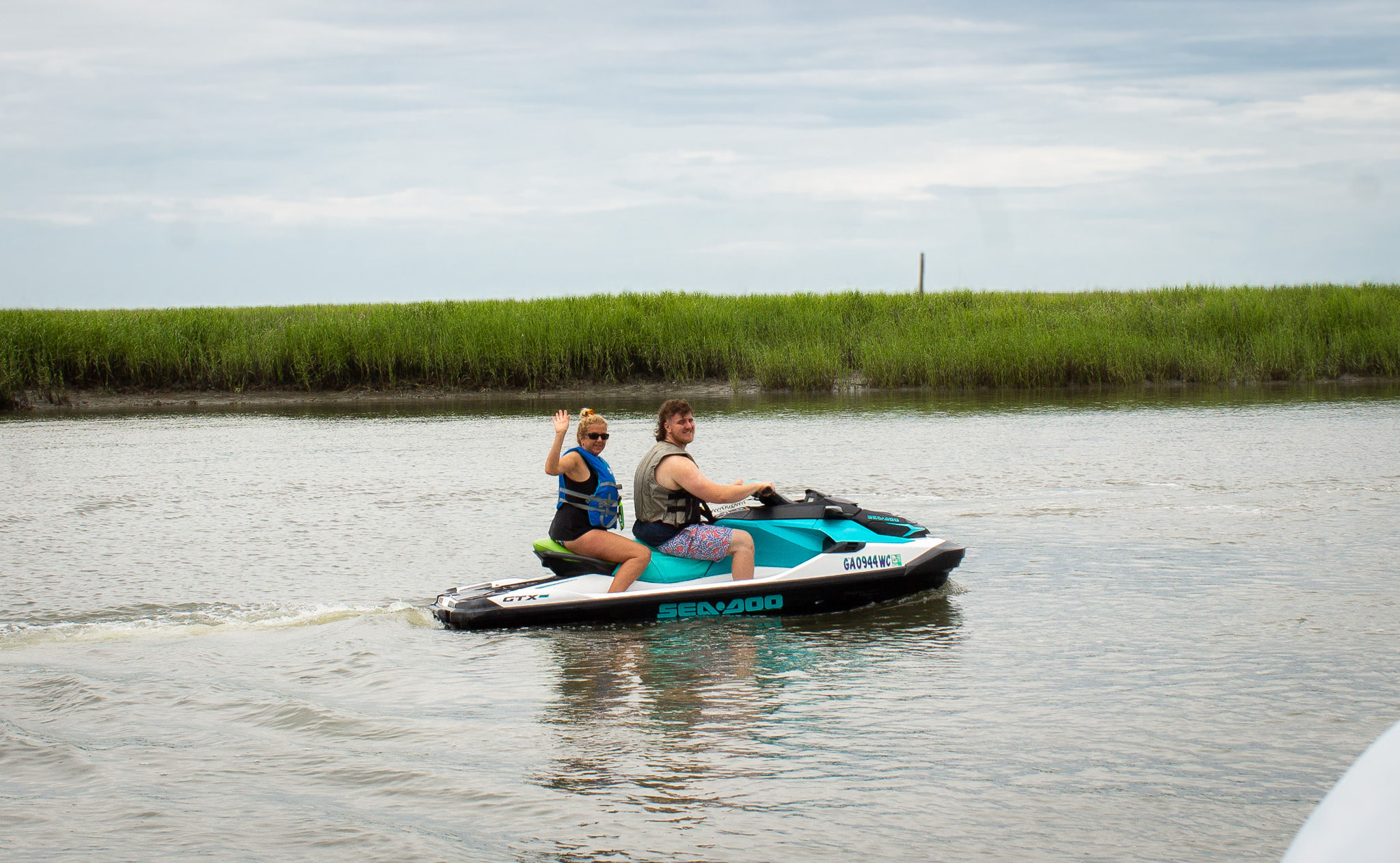 Two people wearing life jackets riding a teal-and-white jet ski through a calm coastal marsh waterway, one passenger waving.