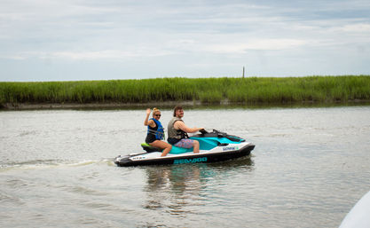 Two people wearing life jackets riding a teal-and-white jet ski through a calm coastal marsh waterway, one passenger waving.