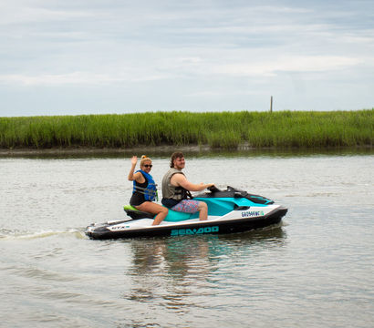 Two people wearing life jackets riding a teal-and-white jet ski through a calm coastal marsh waterway, one passenger waving.
