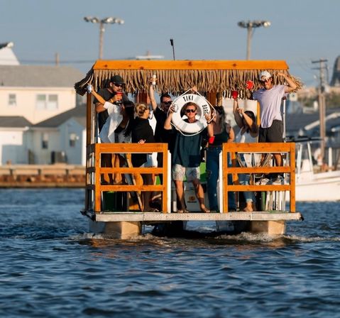 Group of friends partying on a tiki-style pontoon boat with a thatched roof, holding drinks and cheering on a sunny summer afternoon near waterfront homes.