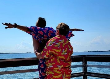 Two people in bright fruit-print shirts (banana and watermelon) pose with arms outstretched on a sunny wooden pier overlooking a calm blue bay and clear sky.