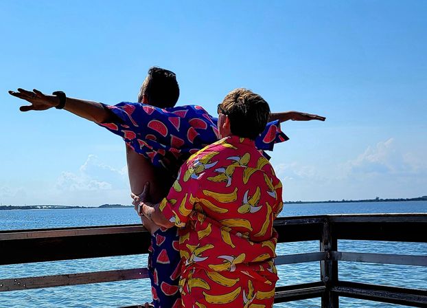 Two people in bright fruit-print shirts (banana and watermelon) pose with arms outstretched on a sunny wooden pier overlooking a calm blue bay and clear sky.