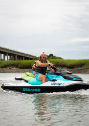 Excited rider in a life vest piloting a turquoise Sea-Doo jet ski with a passenger through shallow coastal Georgia marsh water near a low concrete bridge