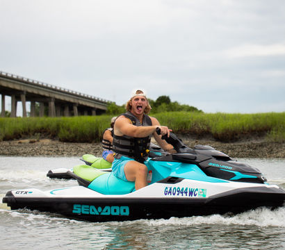 Excited rider in a life vest piloting a turquoise Sea-Doo jet ski with a passenger through shallow coastal Georgia marsh water near a low concrete bridge