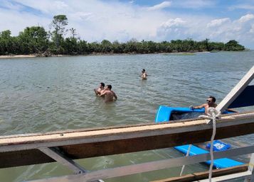 Relaxed scene of three people wading in a shallow coastal bay near a mangrove-lined shore and a fourth reclining on a blue inflatable beside a boat railing under a partly cloudy sky.