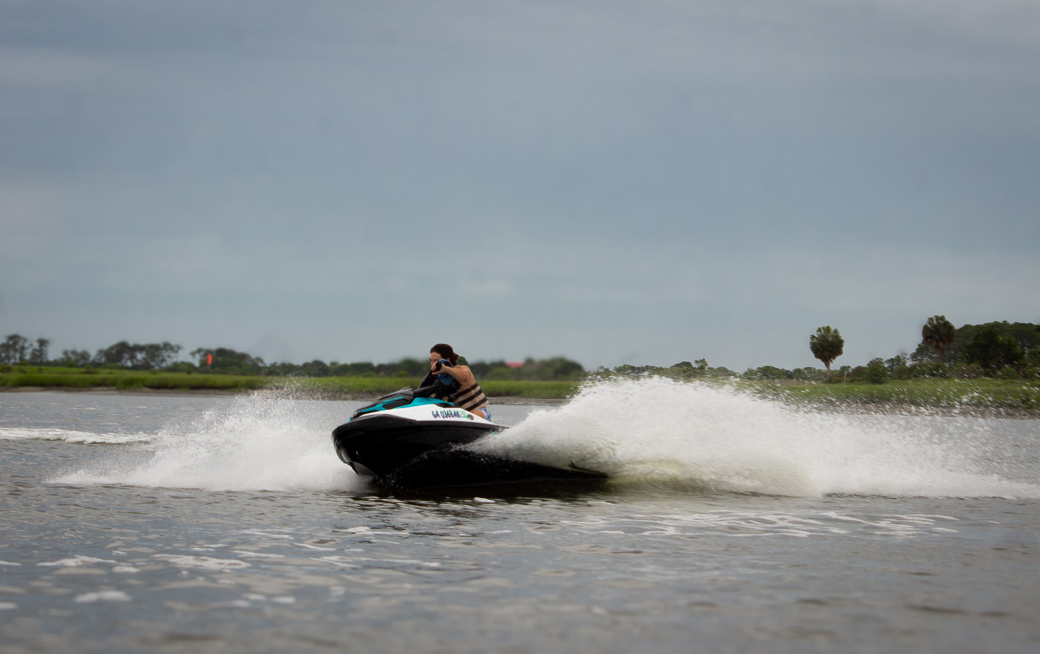 Jet ski speeding across a coastal marsh estuary, rider in a life vest kicking up a large spray of water under a cloudy sky