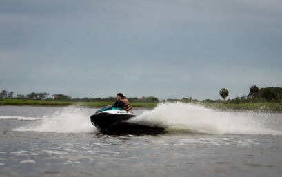Jet ski speeding across a coastal marsh estuary, rider in a life vest kicking up a large spray of water under a cloudy sky