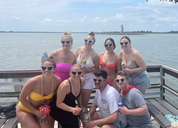 Eight friends in swimsuits wearing heart-shaped sunglasses, holding canned drinks on a wooden dock/boat by a calm bay with a distant lighthouse and sunny sky.