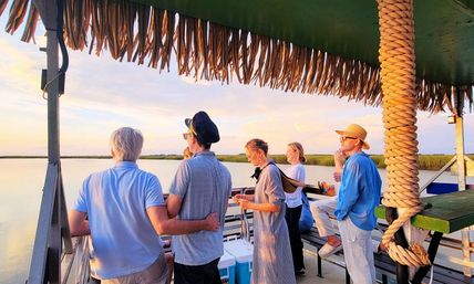 People on a thatched-roof boat deck watching a pastel sunset over calm coastal marsh reeds — relaxed waterfront cruise scene.