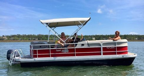 Red-and-white pontoon boat with canopy carrying three smiling adults relaxing on calm coastal waters under a sunny blue sky with a tree-lined shoreline in the background.