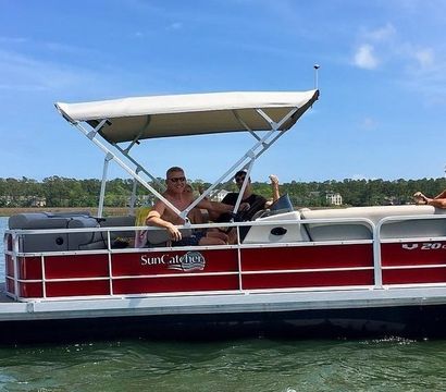 Red-and-white pontoon boat with canopy carrying three smiling adults relaxing on calm coastal waters under a sunny blue sky with a tree-lined shoreline in the background.