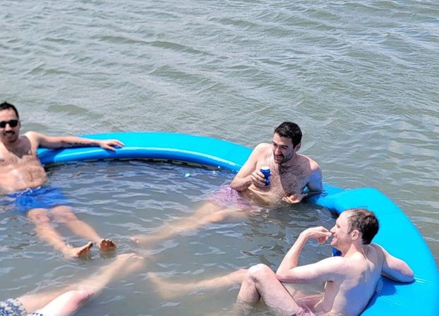 Three men lounging in a round blue inflatable ring on a lake, waist-deep in water and wearing swim trunks—one with sunglasses and another holding a canned drink, chatting and relaxing.