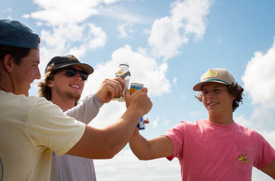 Three friends in caps clinking cans and bottles under a bright blue sky — casual summer cheers outdoors