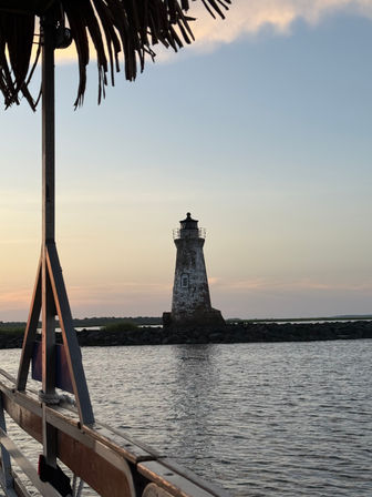 Weathered brick lighthouse on a rocky breakwater at sunset, framed from a boat deck under a thatched canopy with calm coastal waters and marshland background.