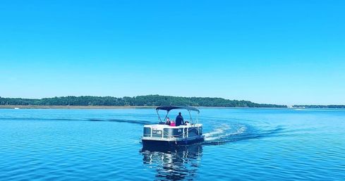 Leisurely pontoon boat with passengers cruising across calm bright-blue water under a clear sky, tree-lined shoreline in the distance.