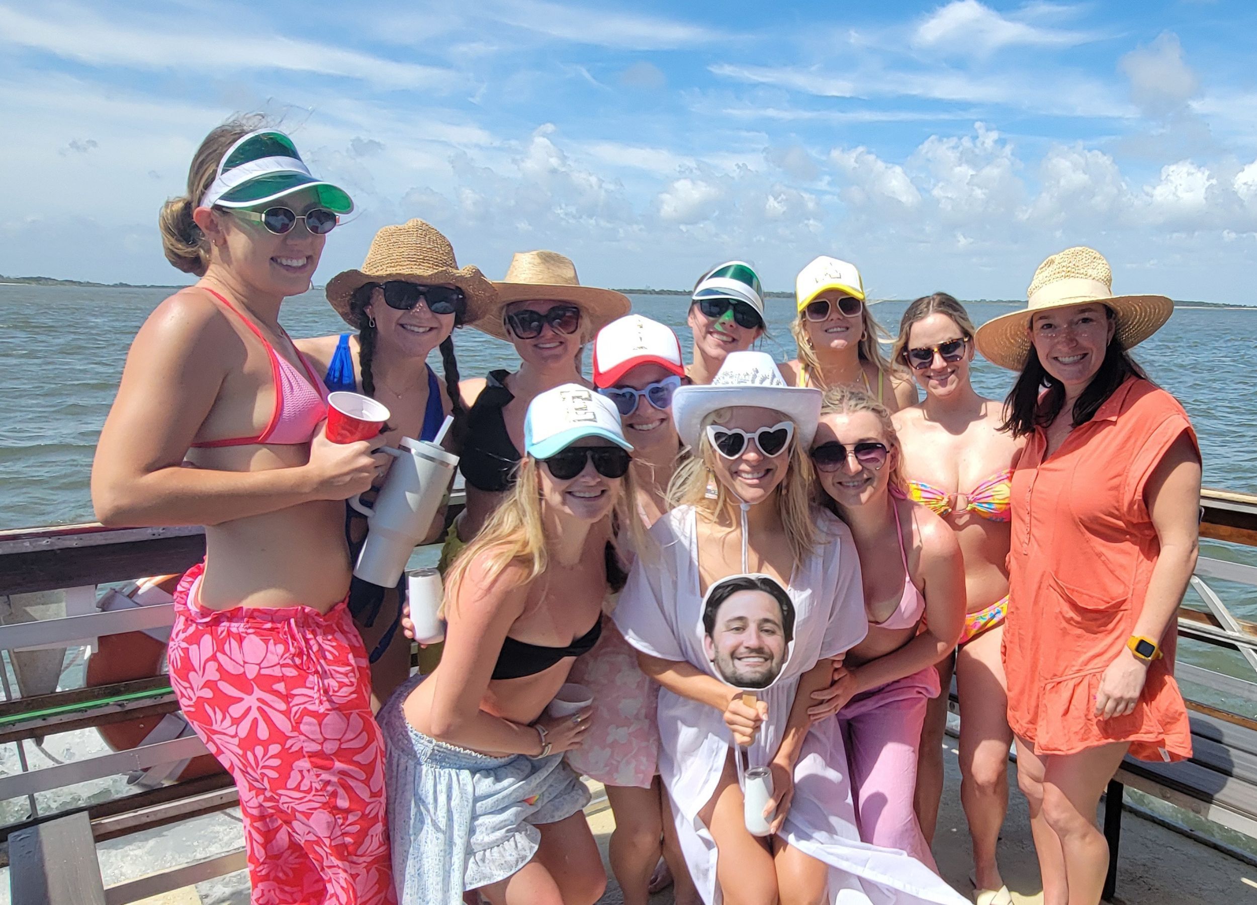 Smiling group of friends in swimsuits and sun hats posing with drinks on a sunny boat over calm coastal waters.